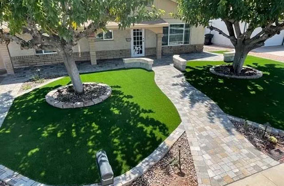 Aerial view of a landscaped yard with two trees and vibrant green artificial grass, bordered by stone pathways and circular flower beds.