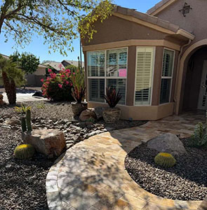 A curved stone pathway leads to a sunlit home entrance, surrounded by desert plants and colorful flowers in a landscaped yard.