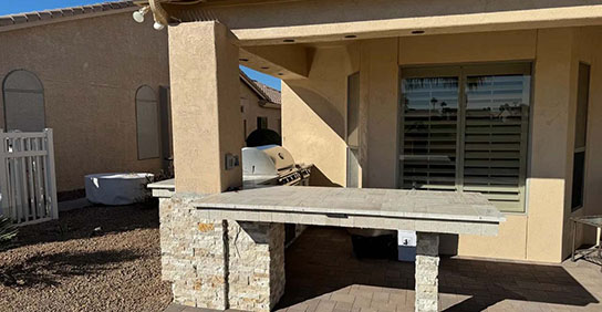 Outdoor patio featuring a stone barbecue area, a grill, and a sturdy table under a shaded roof, surrounded by gravel landscaping.